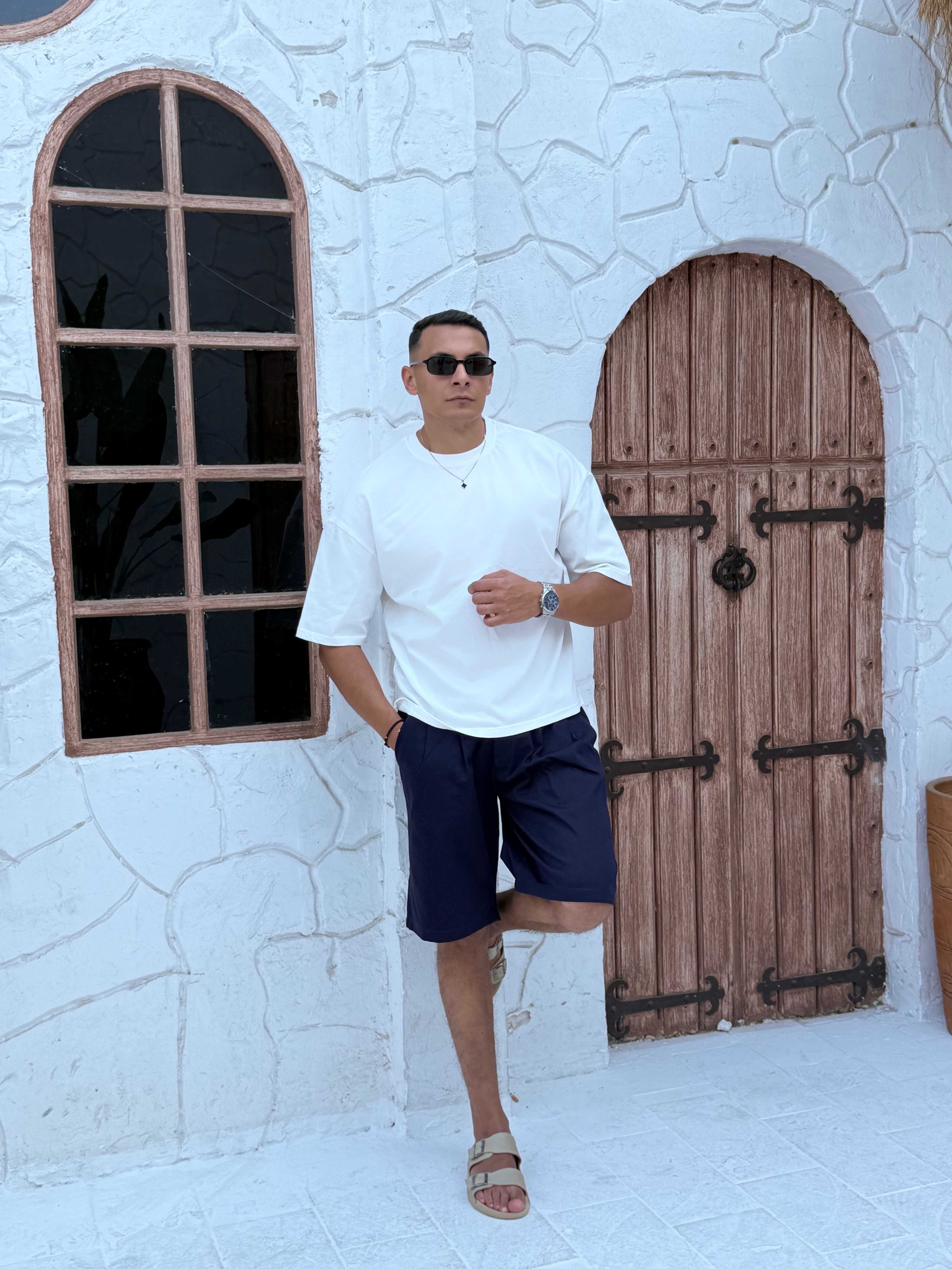 man, wear t-shirt white cotton, fame clothes A man wearing a black cotton t-shirt, white shorts, and beige sandals stands in front of a white textured stucco building with arched windows. From fame clothes 