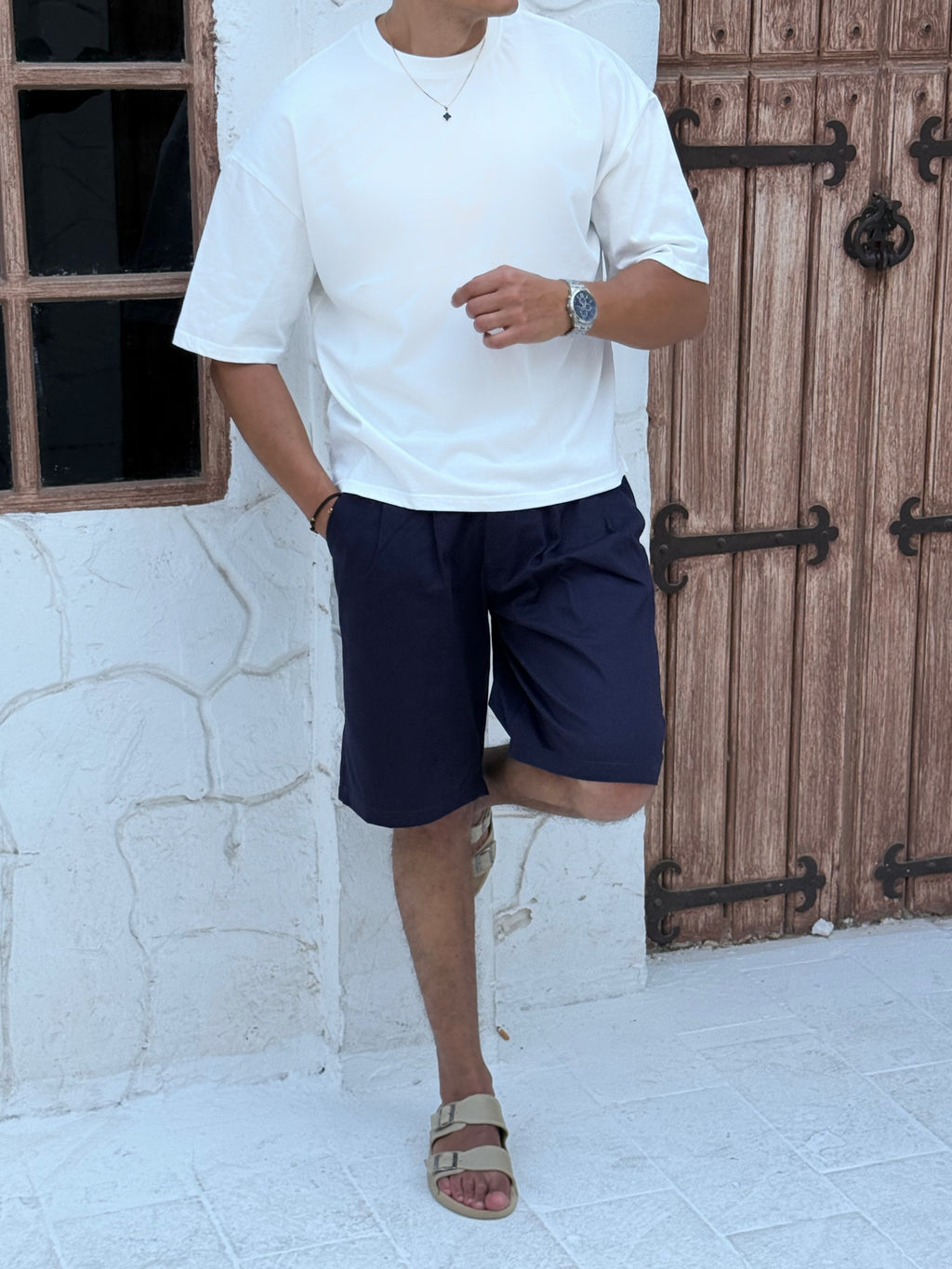 man, wear t-shirt white cotton, fame clothes A man wearing a black cotton t-shirt, white shorts, and beige sandals stands in front of a white textured stucco building with arched windows. From fame clothes 