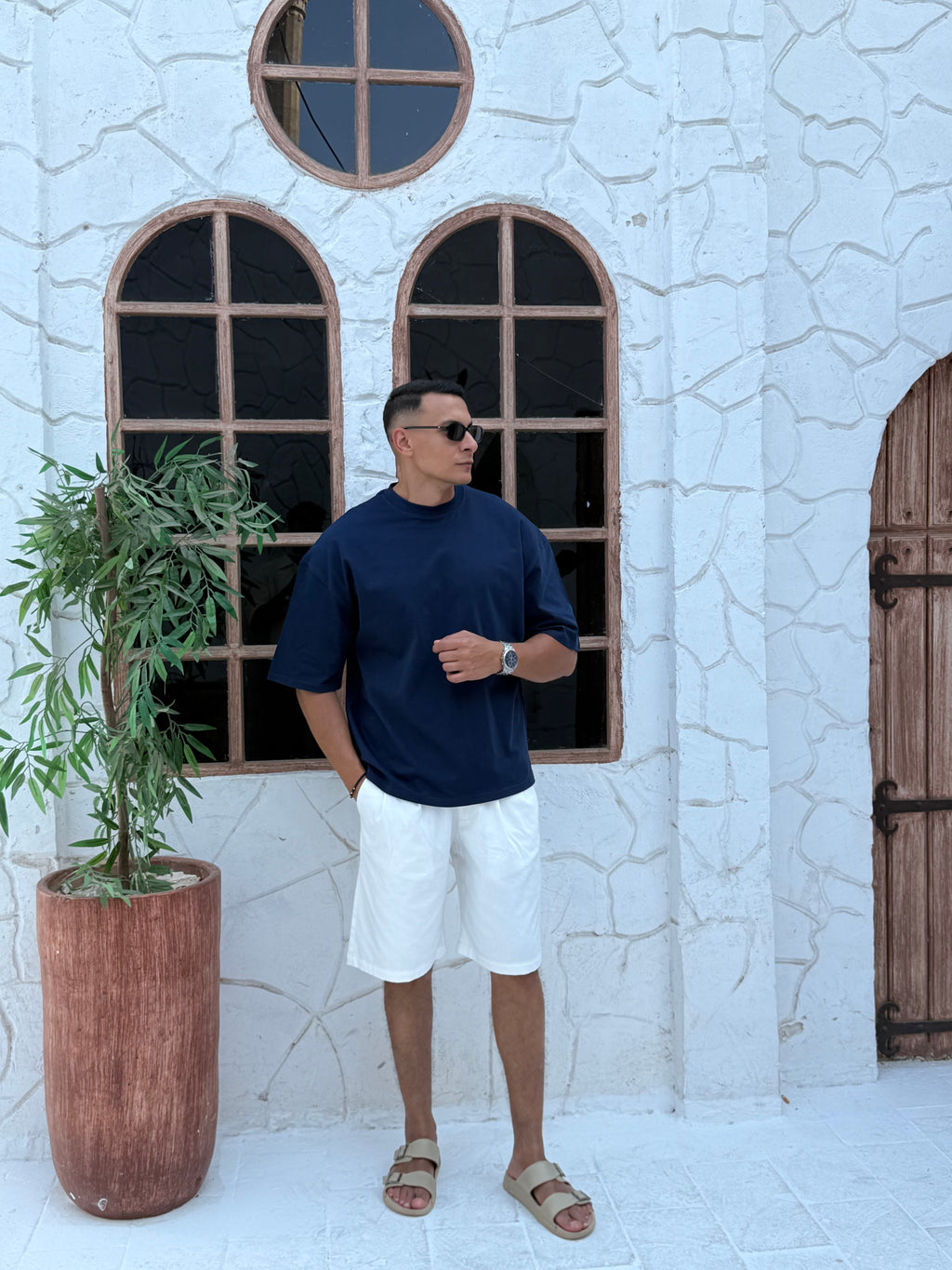 fame clothes A man wearing a navy-blue t-shirt, white shorts, and beige sandals stands in front of a white textured stucco building with arched windows.from fame clothes 
